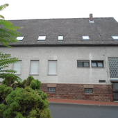 im Vordergrund Straße, links Büsche. Hinten quer stehendes weißes Haus mit grauem Dach. Im 1. OG Fenster, im EG rechts an der Hausecke Eingang.