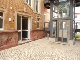 links Teilansicht der aus roten Steinen bestehenden Fassade. In der Mitte Eingang. Neben dem Haus rechts hinten Glasschacht mit Außenaufzug.
