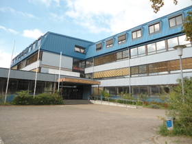 im Vordergrundgepflasterter Platz. L-förmiges Schulgebäude, 3-stöckig. Oben blauer Streifen an der Fassade. Flachdach.