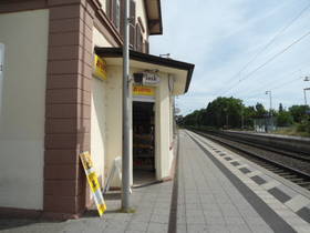 Bahnsteig der S-Bahn. Links Hausecke mit überdachtem Eingang zum Kiosk. Davor links hoher Pfosten. Rechts Sicht auf Bahngleis und Schienen.