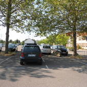 Ein Parkplatz unter Bäumen, mittig parkender PKW. Im Hintergrund Gebäude.