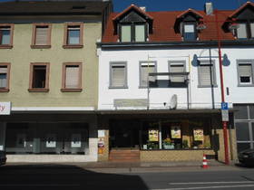 Geäudezeile; rechts weißes 3-stöckiges Haus. Über EG Vordach. Bildmitte: Eingang mit Stufen, rechts großes Schaufenster mit Dekoration und Plakaten.