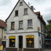 frei stehendes weißes Haus mit rotem Spitzdach. Vor dem Eingang schmale Stufen, links und rechts je 1 Fenster mit je 1 gelben Schild mit Namen.