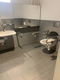 Modern ausgestatteter Toilettenraum mit hellgrauen Fliesen, Weiße Hängetoiletteund weißes Waschbecken, beide  mit Haltegriffen rechts und links ausgestattet. Niedrig aufgehängte Spiegel vorhanden.