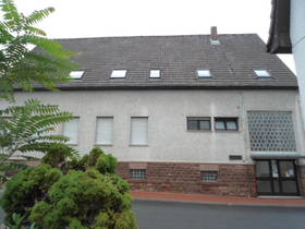 im Vordergrund Straße, links Büsche. Hinten quer stehendes weißes Haus mit grauem Dach. Im 1. OG Fenster, im EG rechts an der Hausecke Eingang.