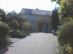 Bildmitte gepflasterter Zugangsweg durch Garten, im Hintergrund 2-stöckiges helles Haus.