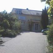 Bildmitte gepflasterter Zugangsweg durch Garten, im Hintergrund 2-stöckiges helles Haus.