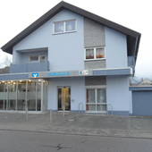 2-stöckiges hellblaues Haus. Frontansicht von Straße her. Im EG links Eingang zur Bank. Vor dem Haus Gehweg und Straße.g