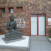 rotes Backsteinhaus. links durch Randsteine abgesetztes Beet mit weißen Steinen, in dem große Buddha-Figur steht. daneben rechts kleiner Weg bis zur Türe mit weißen Jalousien.