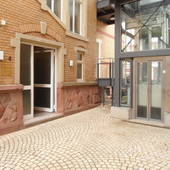 Links Teilansicht Backsteinhaus, mittig Eingangstür. Rechts am Haus angrenzender Anbau des Glasaufzuggschachtes.