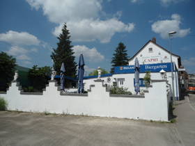 weiße, halbhohe Mauer vor der Terrasse, dahinter Sitzbreich mit blauen Sonnenschirmen nur teilweise sichtbar. Im Hintergrund weißes Haus mit blauem Poster "Biergarten" und Namenslogo "Capri".