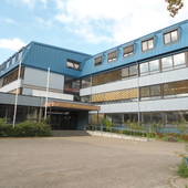 im Vordergrundgepflasterter Platz. L-förmiges Schulgebäude, 3-stöckig. Oben blauer Streifen an der Fassade. Flachdach.