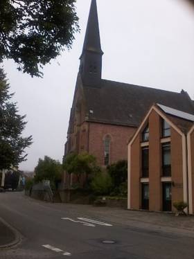 Ansicht der Kirche von der Straße her. Rechts Gebäude mit Kirchturm links. Daneben links ansteigende Straße.