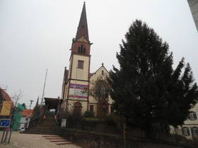 Im Hintergrund Kirche mit Kirchturm auf Anhöhe. Links Treppe, rechts Hangbepflanzung mit Bäumen und Büschen.