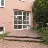 rosafarbenes Gebäude. Mittig Eingang, davor 2 breite Stufen. Rechte Bildseite Büsche.
