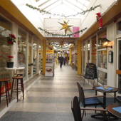 breiter Innenflur, beidseits Ladengeschäfte, im Vordergrund rechts blaue Stühle und Tisch eines Cafes. Glasdach über Passage.hts