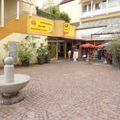 links Teilansicht des gellben Gebäudeteils, dort an der Hausecke Eingang zur Bar. Auf dem Platz links Steinbrunnen. Hinten quer und rechts Geschäfte.