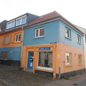 Halbseitenansicht des Hauses: EG rosa mit blauer Tür, davor 2 Stufen. 1. OG hellblau. Hausanteil links mit spiegelverkehrter Farbgebung. Daneben rechts weißes Haus. Vorne Teil des Gehwegs.
