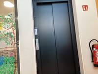 Aufzug mit dunkler geschlossener Tür in heller Wand.
Rechts neben Aufzug hängt ein Feuerlöscher an der Wand.