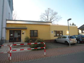 Im Hintergrund Eingangsbereich. Gelber Flachbau, links Glastür, rechts Fenster. Vor dem Gebäude gepflasterter kleiner Platz.