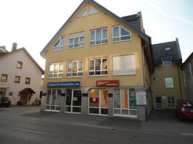 gelbes freistehendes 3-stöckiges Haus. Im EG Fensterfront. Eingang zu Geberth um die Hausecke links. Im Hintergrund links Wohnhaus.