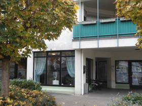weißes mehrstöckiges Haus, großes Schaufenster im EG, daneben rechts Eingangsbereich: links Tür zum Nagelstudio, 2 Stühle und kleiner Tisch neben der Tür.