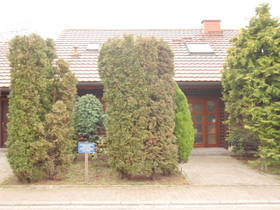Wohnhaus mit Holzfassade. Am Gehweg links 2 hohe Strächer, daneben rechts schmaler Durchang  zur Haustür. Neben der Tür rechts Strauch.