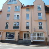 4-stöckiges rosa Haus. Links Einfahrt, danben rechts Schausfenster, mittig Treppe, oben rechts Eingang zur Eisdiele. Daneben rechts Schaufenster mit weißen Sprossen. Vor dem Gebäude Teilansicht der Straße.
