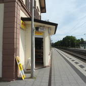 Bahnsteig der S-Bahn. Links Hausecke mit überdachtem Eingang zum Kiosk. Davor links hoher Pfosten. Rechts Sicht auf Bahngleis und Schienen.