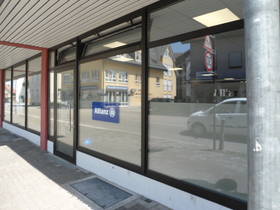 Im EG mehrere große Schaufenster und Glastüren in einer Reihe. Im rechten Bildbereich Eingangstüre zum Büro. im