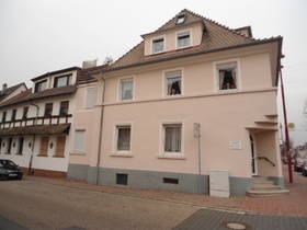 3-stöckiges rosa Eckhaus. Spitzdach mit Gaubenfenstern. In der Hausecke Eingang zur Praxis, davor Stufen.