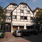 Fachwerkhaus mit Spitzdach. Im EG mit kleiner Markise überdachte Schaufenster, in der Mitte Eingangstür. Am Bildrand links und rechts Bäume.