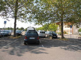 Ein Parkplatz unter Bäumen, mittig parkender PKW. Im Hintergrund Gebäude.